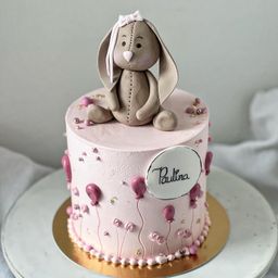 Ein rosa Geburtstagskuchen mit einer Hasenfigur oben drauf, geschmückt mit rosa Luftballons und Blumen, steht auf einem goldenen Teller. Der Kuchen hat eine zarte Glasur und eine persönliche Nachricht für Paulina.