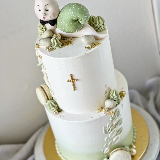 Ein zweistöckiger weißer Kuchen mit grünen und goldenen Dekorationen zeigt eine verspielte Hasenfigur und ein goldenes Kreuz auf der mittleren Etage.