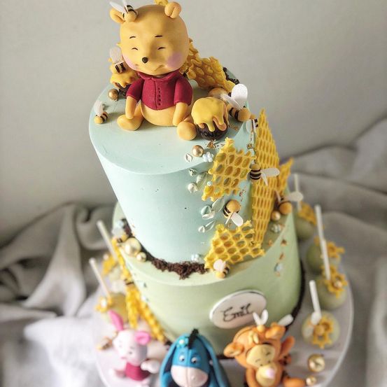 Ein zweistöckiger Kuchen mit Winnie Puuh obenauf, umgeben von Bienen und Honigwaben-Dekorationen, und anderen Charakteren wie I-Aah und Tigger im unteren Stockwerk.