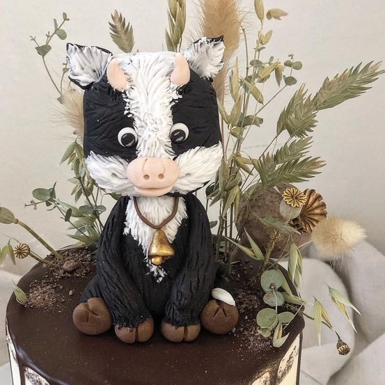 Ein Schokoladenkuchen, verziert mit einer Figur einer schwarz-weißen Kuh mit einer Glocke um den Hals, umgeben von verschiedenen grünen Pflanzen und Blumen.