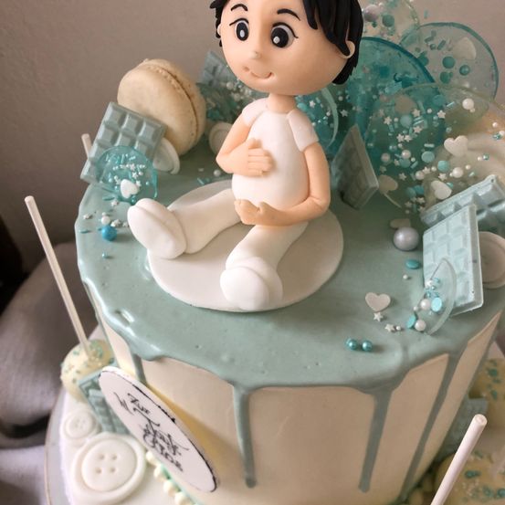 Ein Kuchen mit einer Figur eines Jungen oben drauf, umgeben von blauen Dekorationen, einem Macaron und einem Schild, das 'Es ist ein Junge' sagt.