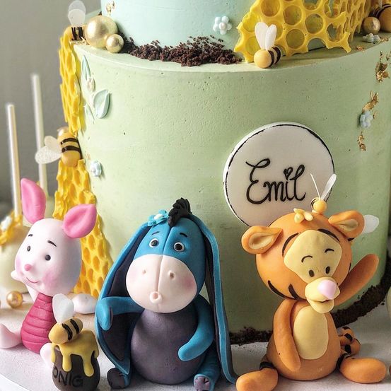 Ein bunter Kuchen zeigt Winnie Puuh, I-Ah und Tigger mit einer grünen Glasur und Wabendesign. Der Kuchen trägt den Namen Emil in der Mitte und kleine Figuren von Winnie Puuh und einer Biene.