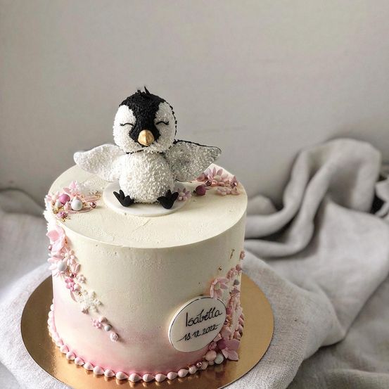 Ein weißer Kuchen mit einem Pinguin oben drauf, dekoriert mit rosa Blumen und Perlen. Der Kuchen hat ein Etikett mit dem Namen Isabella und dem Datum 18.12.2022.