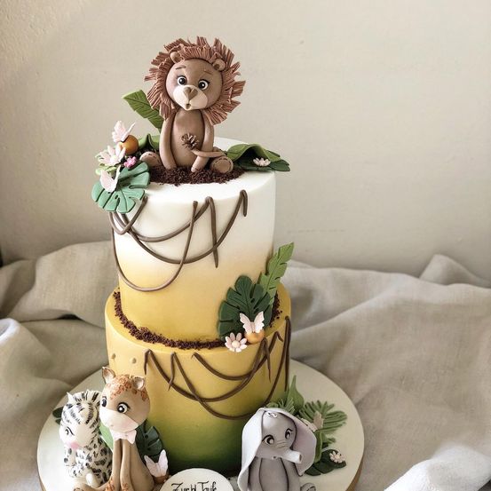 Ein dreistöckiger Kuchen mit einem Löwen oben, umgeben von Dschungelelementen. Unten sind ein Tiger und ein Giraffen vorhanden. Der Kuchen hat ein gelbes und weißes Farbschema.