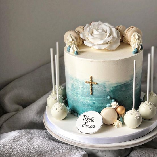 Ein Kuchen mit einer weißen Rose und einem goldenen Kreuz oben, umgeben von weißen Macarons und Cake Pops. Der Kuchen hat einen blau-weißen Farbverlauf.