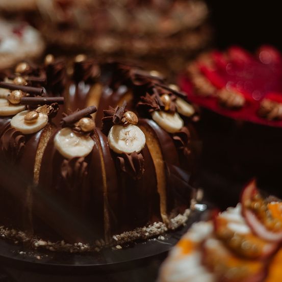 Ein Nahaufnahme eines schokoladenüberzogenen Gugelhupfs mit geschnittenen Bananen und Schokoladenraspeln. Andere Desserts sind im Hintergrund verschwommen.