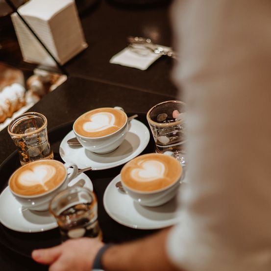 Eine Person hält einen Tablett mit drei Kaffeetassen, jede mit einem Herzdesign, zusammen mit Gläsern und Untertassen auf einem Holztisch mit unscharfem Hintergrund.