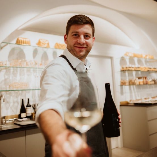 Ein lächelnder Mann in einem weißen Hemd und einer Schürze hält eine Flasche Wein und ein Weinglas in einer Küche. Hinter ihm befinden sich Regale mit Backwaren und Weinflaschen.