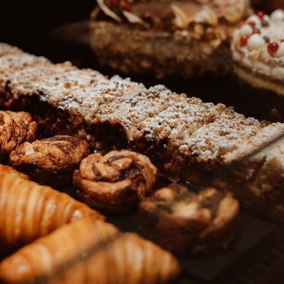 Eine Auswahl an Gebäck, darunter ein langes Gebäck mit Puderzucker, mehrere Zimtschnecken und Croissants, ist in einer Bäckerei-Vitrine ausgestellt.