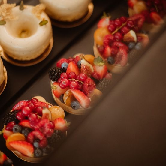 Eine Nahaufnahme einer Display von Mini-Gebäckstücken, einige gefüllt mit Früchten und andere mit Sahne. Die Früchte umfassen Beeren, Erdbeeren und andere verschiedene Früchte.