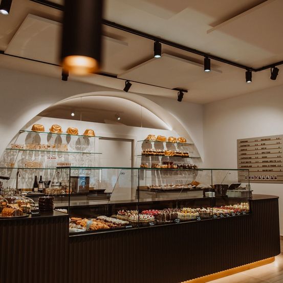 Eine Bäckerei mit modernem Design, die eine Glastheke mit einer Auswahl an Gebäck wie Croissants und Kuchen präsentiert. Die Regale sind mit verschiedenen Backwaren gefüllt. Oben beleuchten Schienenleuchten den Raum.