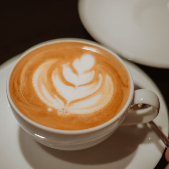 Ein Nahaufnahme einer weißen Keramik-Kaffeetasse mit einem Latte-Art-Design auf einer Untertasse. Der Kaffee ist eine Mischung aus Braun und Weiß.