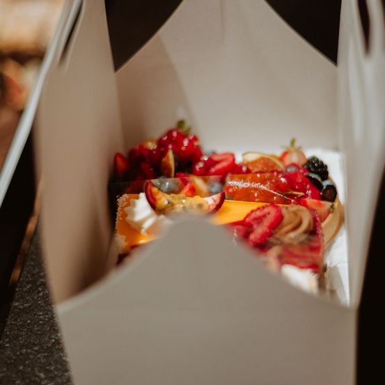 Eine weiße Box mit Deckel zeigt eine Vielfalt an Fruchttörtchen, darunter Erdbeeren, Brombeeren und Orangenscheiben, ordentlich im Inneren angeordnet.