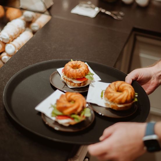 Eine Person hält einen Tablett mit drei Sandwiches. Jedes Sandwich befindet sich auf einem Teller mit einer Serviette. Im Hintergrund sind Gebäckstücke zu sehen.