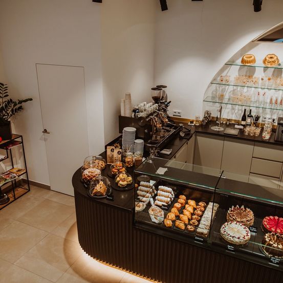 Eine moderne Bäckerei mit einem Tresen, der verschiedene Gebäcke, Kuchen und Brote anbietet. Der Tresen besteht aus Glas und Holz, mit Regalen darüber, die Gläser und Flaschen halten. Eine Kaffeemaschine steht auf dem Tresen, und eine Pflanze befindet sich auf einem nahen Regal.