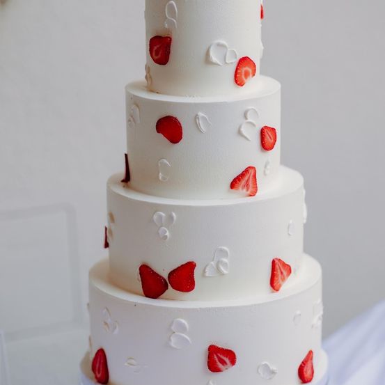 Ein vierstöckiger weißer Kuchen, verziert mit herzförmiger weißer Glasur und roten Erdbeeren. Jede Etage hat eine einzigartige Erdbeer-Arrangement.