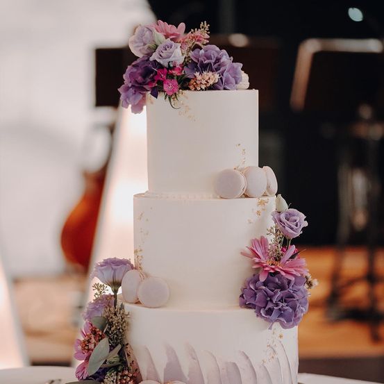 Ein dreistufiger weißer Kuchen mit Goldakzenten, dekoriert mit lila und rosa Blumen und Macarons. Der Kuchen steht auf einem Tisch, mit Stühlen und einem unscharfen Hintergrund.