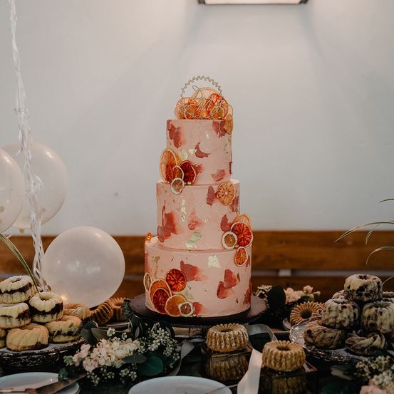 Ein großer mehrstöckiger Kuchen mit rosa Glasur und Orangenscheiben. Er steht auf einem Tisch mit Luftballons, Blumen und anderen Desserts.