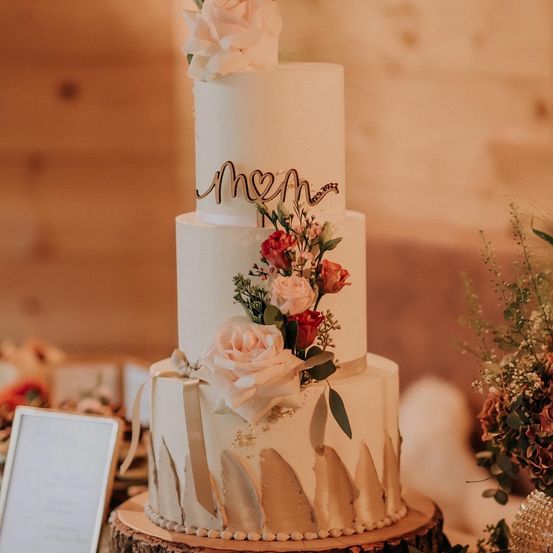 Eine dreistöckige weiße Hochzeitstorte, verziert mit weißen und rosa Rosen, grünen Blättern und einer herzförmigen goldenen Inschrift. Sie steht auf einem Holzsockel, mit einem unscharfen Hintergrund einer Holzwand.