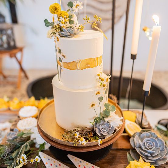 Ein dreistöckiger weißer Kuchen mit Zitronen- und Gänseblümchendekorationen steht auf einer goldenen Platte. Er ist umgeben von Sukkulenten, Zitronen und Kerzen auf einem Holztisch.
