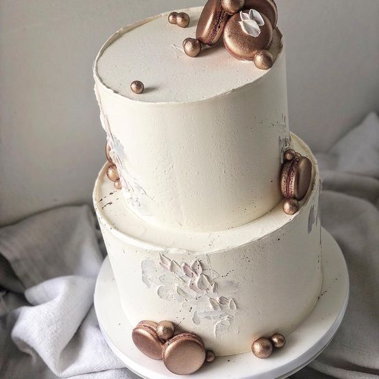 Ein dreistufiger weißer Kuchen mit bronzefarbenen Akzenten und einem zarten weißen Blumendesign. Er hat einen glänzenden Finish und ist mit bronzefarbenen Macarons gekrönt.