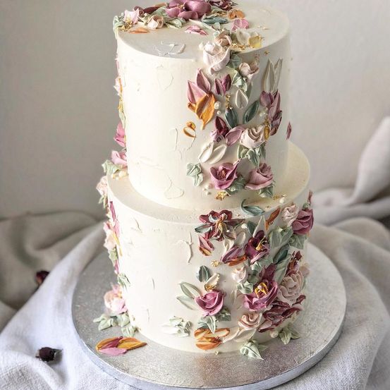 Ein dreistufiger weißer Kuchen mit komplizierten Blumendesigns, einschließlich Rosen, Blättern und Blütenblättern, auf einem silbernen Teller.