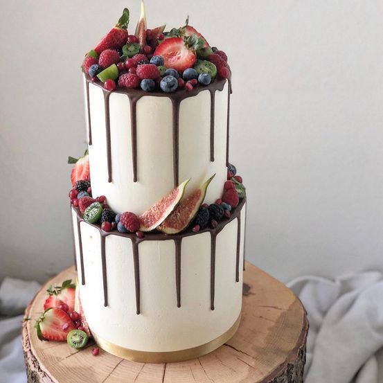 Ein dreistöckiger Kuchen mit weißer Glasur, Schokoladensauce und einer Vielzahl frischer Beeren und Feigen darauf, präsentiert auf einer Holzplattform.