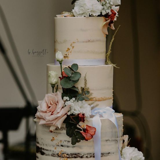 Ein dreistöckiger Hochzeitskuchen mit weißer und goldener Glasur, geschmückt mit weißen, rosa und roten Blumen sowie grünen Blättern. Ein Band ist um die mittlere Etage gebunden.
