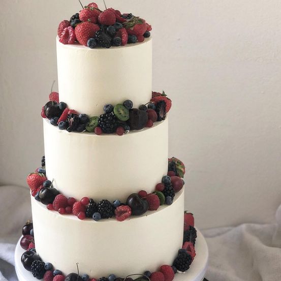 Ein weißer dreistöckiger Kuchen mit einer Vielfalt von Beeren obenauf, darunter Erdbeeren, Blaubeeren, Himbeeren und Brombeeren. Der Kuchen steht auf einer weißen Oberfläche.