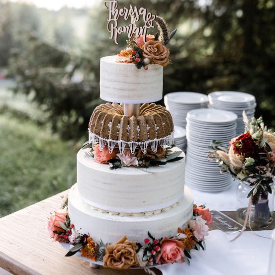 Ein dreistufiger Hochzeitskuchen mit Blumenschmuck steht auf einem Holztisch, neben Stapeln von Tellern und einem Blumenstrauß. Der Kuchen hat oben ein Bundt-Design mit einem dekorativen Schild, das 'Theresa & Roman' steht.