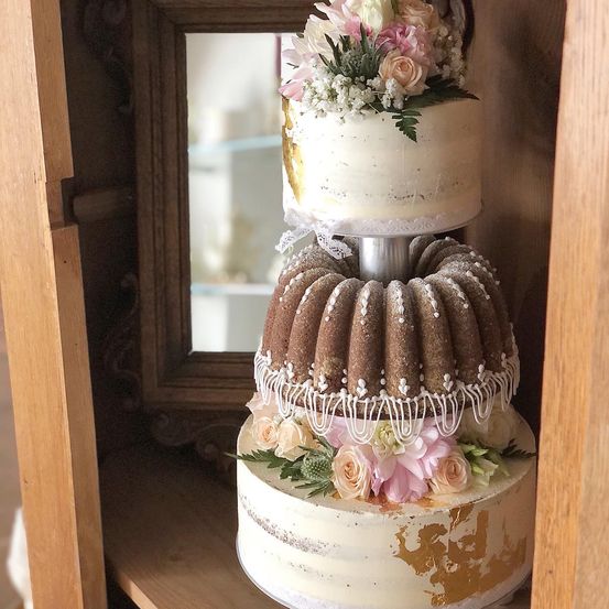 Ein dreistöckiger Kuchen mit einer Bundt-Kuchen-Mittelschicht, verziert mit rosa und weißen Blumen, wird auf einem Holzregal präsentiert.