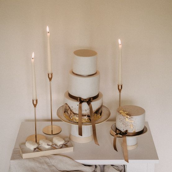 Drei elegante weiße und goldverzierte Kuchen sind auf einem Tisch mit drei brennenden Kerzen auf Messingkerzenständern ausgestellt. Ein Teller hält ein kleines Tablett mit weißen Dessert-Leckereien.