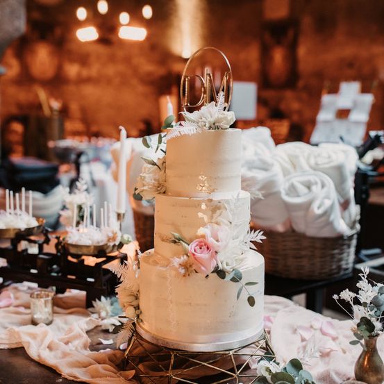 Ein dreistöckiger Hochzeitskuchen, verziert mit weißen Blumen und Grün, steht auf einem Tisch. Dahinter ist ein Korb mit gefalteten weißen Handtüchern zu sehen. Der Kuchen hat einen monogrammierten Topper und ist von Kerzen und anderen Dekorationen umgeben.