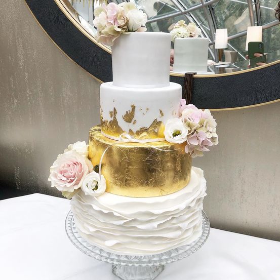 Ein dreistöckiger Kuchen mit Gold- und Weißglasur. Die Spitze hat eine goldene Schicht mit einem Goldblattdesign. Rosa Blumen schmücken die Seiten.