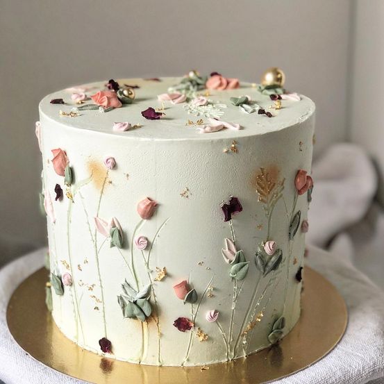 Ein weißer Kuchen mit Blumendesign ist mit verschiedenen Blumen und Blättern verziert. Er steht auf einem goldenen Teller mit einem weißen Tuch darunter.
