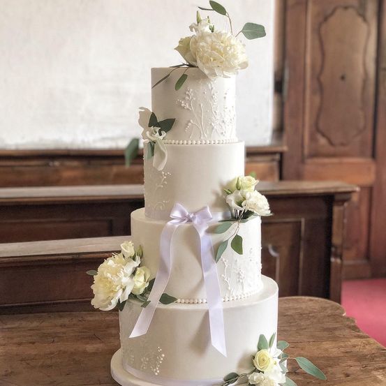 Ein dreistöckiger weißer Hochzeitskuchen, geschmückt mit weißen Blumen und grünen Blättern, auf einem Holztisch. Im Hintergrund sind hölzerne Kirchenbänke zu sehen.