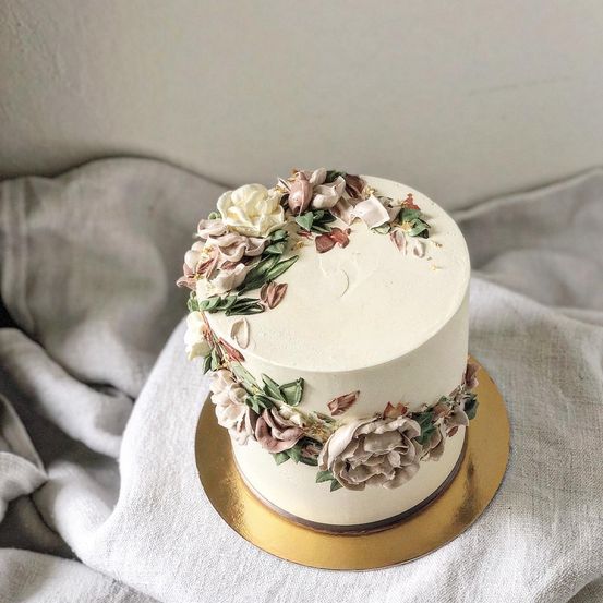 Ein weißer Kuchen mit dekorativen Blumenmustern steht auf einem goldenen Kuchenständer. Der Kuchen ist mit weißen und rosa Blumen verziert, und um ihn herum sind kunstvoll grüne Blätter angeordnet.
