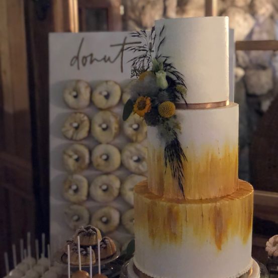 Ein dreistöckiger Kuchen mit weißer Glasur und einem Goldguss auf den unteren beiden Etagen. Er ist mit Blumen dekoriert und ein Schild mit dem Wort 'Donut' im Hintergrund.