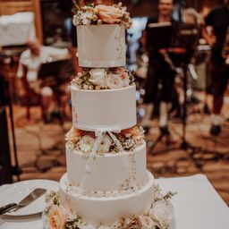 Ein vierstöckiger weißer Hochzeitskuchen mit Blumenarrangements und Bändern steht auf einem Tisch. Dahinter spielen Musiker in einem unscharfen Hintergrund.