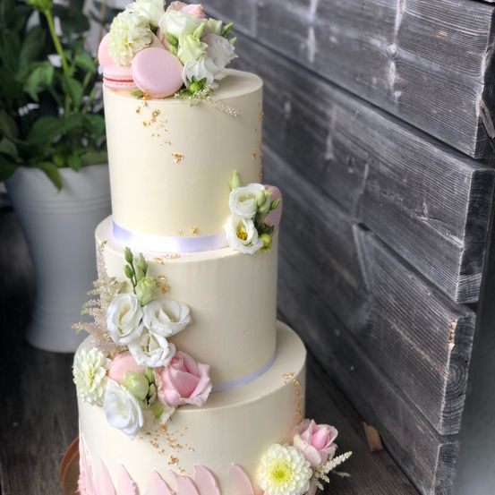 Ein dreistöckiger weißer Kuchen mit rosa und weißen Blumen, rosa Macarons und goldenen Details steht auf einer Holzoberfläche. Ein Topf steht hinter dem Kuchen.