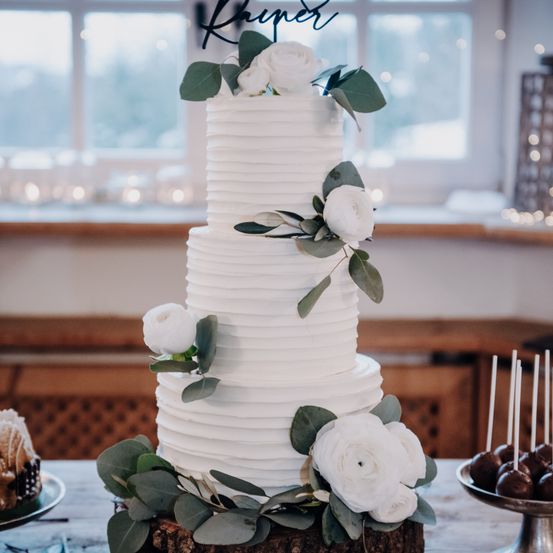 Ein dreistöckiger weißer Hochzeitskuchen mit glatter Glasur, weißen Blumen und grünen Blättern steht auf einem Holzstumpf, mit Schokoladentrüffeln und anderen Desserts in der Nähe.