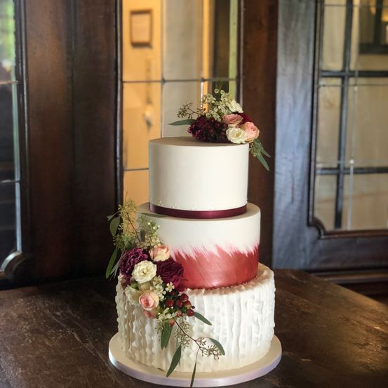 Ein dreistöckiger Hochzeitskuchen mit weißem Fondant und roten Akzenten, geschmückt mit Blumenarrangements, steht auf einem Holztisch.