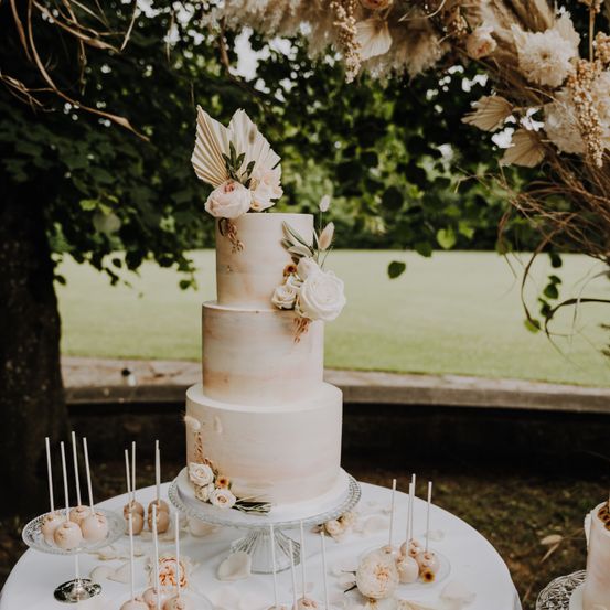 Ein dreistöckiger Hochzeitskuchen mit pastellrosa Glasur und Blumendekorationen steht auf einem Tisch, umgeben von Lutschern und kleinen Kuchen, in einem Garten platziert.