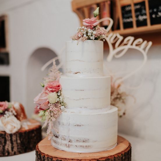 Ein dreistufiger weißer Hochzeitskuchen mit marmorierter Glasur, geschmückt mit rosa und weißen Blumen, steht auf einem Holzstamm. Im Hintergrund zeigt ein Holzregal verschiedene Gegenstände.