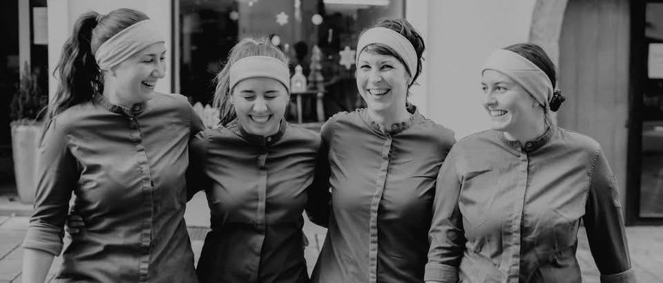 Fünf Frauen stehen lächelnd zusammen draußen. Sie tragen gleiche graue Uniformen mit weißen Stirnbändern und schwarzen Hosen. Sie scheinen für ein Foto zu posieren.