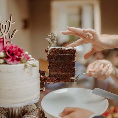 Eine Person schneidet ein Stück Schokoladenkuchen mit einem Messer. Der Kuchen hat eine weiße Glasur und eine Blume darauf. Ein Teller und ein Messer sind auf der rechten Seite. Eine andere Person hält einen Kuchen mit einem Holzschild.