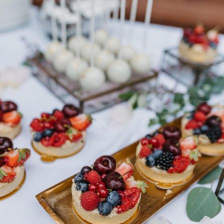 Ein Tisch ist mit einer Auswahl an Mini-Desserts geschmückt. Im Vordergrund befinden sich Törtchen, die mit einer Mischung aus Erdbeeren, Himbeeren und Kirschen belegt sind. Dahinter steht ein Tablett mit weiß glasierten Cake-Pops.