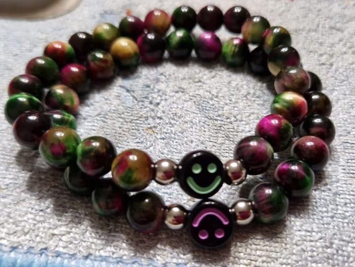 Ein Nahaufnahme eines Armbands mit bunten Perlen. Es enthält mehrere Perlen in verschiedenen Grün-, Lila- und Rottönen. Das Armband hat auch Smiley-Gesichtsperlen.