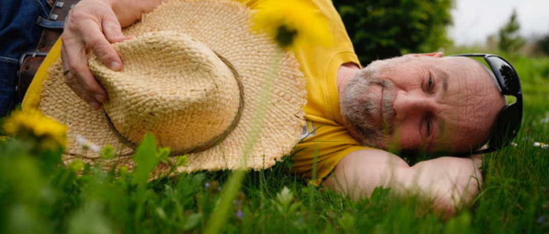 A man with a straw hat lies on the grass, looking upwards with a smile.