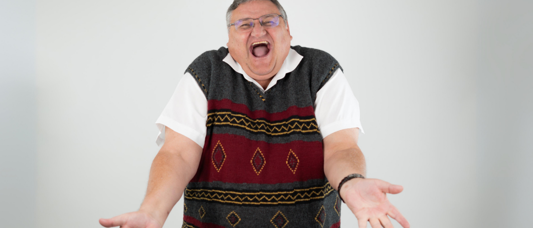 A man wearing glasses and a sweater vest is laughing with his mouth open and hands wide open.